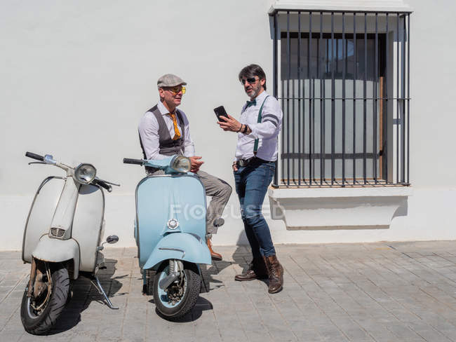 Hipsters positivos de mediana edad en ropa elegante con motos retro tomando una selfie en un día soleado - foto de stock