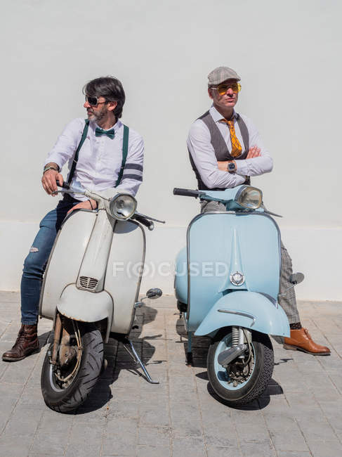Positive middle-aged hipsters in elegant clothes with retro motorbikes looking away in sunny day — Stock Photo