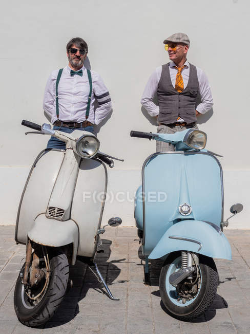 Des hipsters rêveurs d'âge moyen vêtus de vêtements à la mode avec des motos rétro penchées sur le mur par une journée ensoleillée — Photo de stock