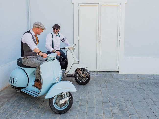 Hipsters positivos de mediana edad en ropa elegante con motos retro mirándose en un día soleado - foto de stock