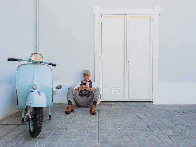 Positive middle-aged hipster in elegant clothes near retro motorbike sitting on the floor and using smartphone on a sunny day — Stock Photo