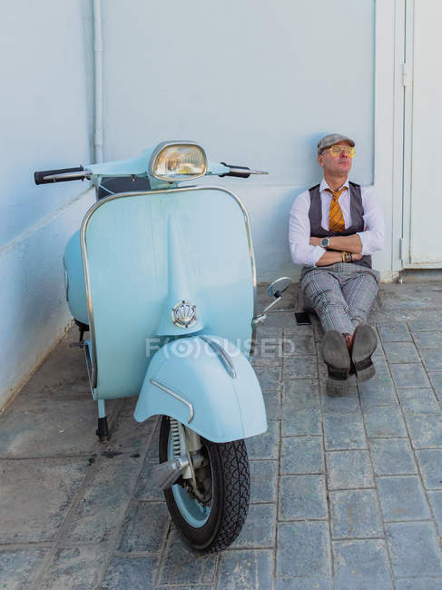 Positive middle-aged hipster in elegant clothes near retro motorbike resting while sitting on the floor on a sunny day — Stock Photo