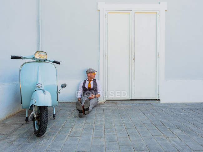 Положительный хипстер средних лет в элегантной одежде возле ретро мотоцикла отдыхает, сидя на полу в солнечный день — стоковое фото