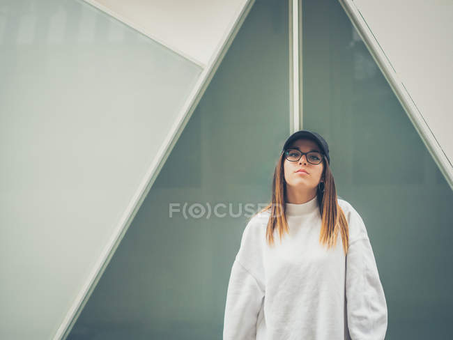 Joven hipster femenino moderno en ropa casual en el fondo de la pared geométrica mirando a la cámara - foto de stock