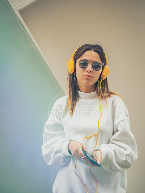 Femme hipster moderne confiante dans des vêtements décontractés en utilisant des écouteurs jaune vif et smartphone sur fond de mur — Photo de stock