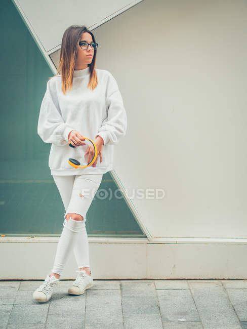 Jovem hipster feminino moderno sonhador em roupas brancas casuais segurando fones de ouvido amarelo brilhante no fundo da parede — Fotografia de Stock