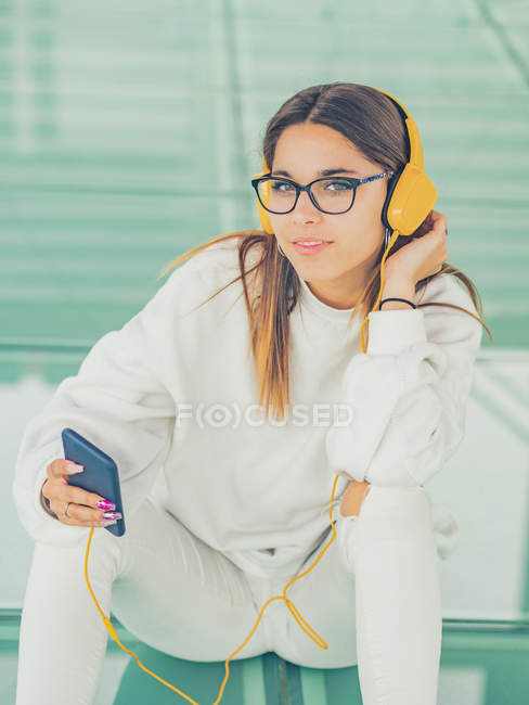 Jeune hipster féminine moderne en vêtements décontractés sur fond de lunettes géométriques écoutant de la musique avec smartphone et regardant la caméra — Photo de stock