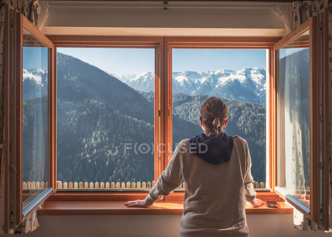 Donna irriconoscibile in abito casual appoggiata sul davanzale della finestra e ammirando cresta verde di montagna nella giornata di sole in campagna — Foto stock