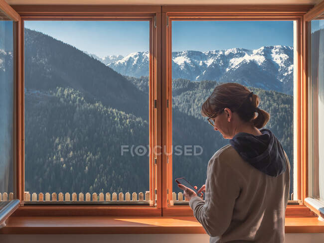 Mujer navegando teléfono inteligente cerca de ventana - foto de stock