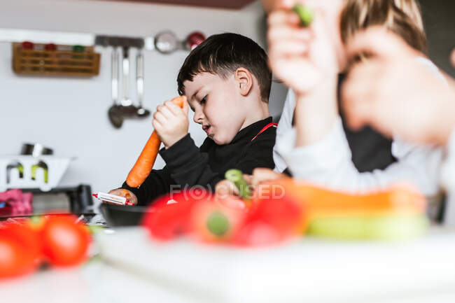 Niñas y niños cortando y pelando verduras maduras mientras cocinan ensalada saludable en la cocina juntos - foto de stock