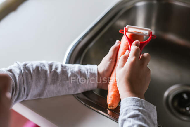 Vista en primera persona del niño anónimo pelando zanahoria mientras cocina ensalada saludable en la cocina - foto de stock