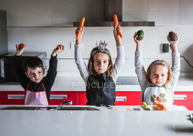Little girls and boy in aprons holding various fresh vegetables in raised hands and looking at camera while standing in kitchen at home together — Stock Photo