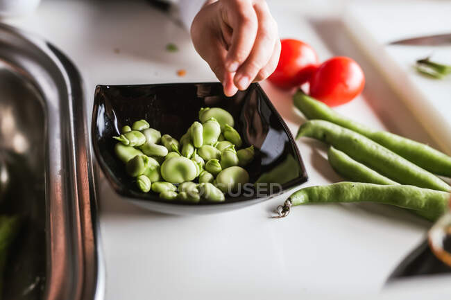 Рука анонімної маленької дівчинки, яка чистить стиглі боби, готуючи здоровий салат на кухні — стокове фото