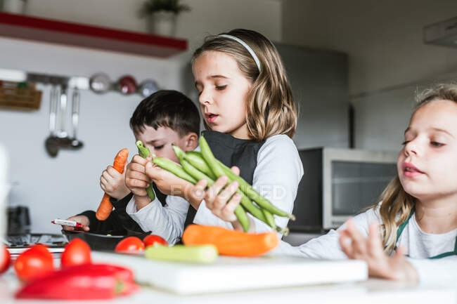 Маленькі дівчата і хлопчик ріжуть і очищають стиглі овочі, готуючи здоровий салат на кухні разом — стокове фото