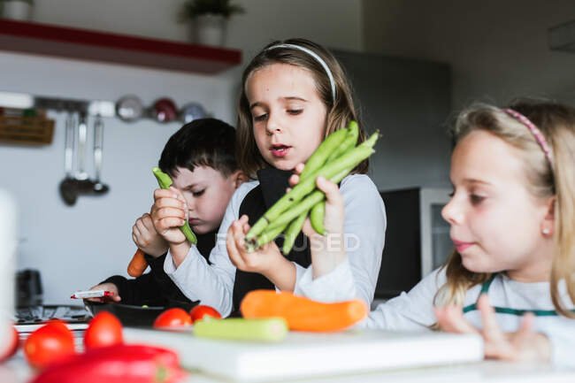 Маленькі дівчата і хлопчик зрізають і чистять стиглі овочі, готуючи здорові салати на кухні разом — стокове фото