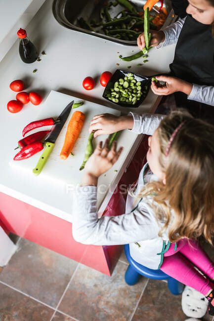 Meninas e meninos cortando e descascando legumes maduros enquanto cozinham salada saudável na cozinha juntos — Fotografia de Stock