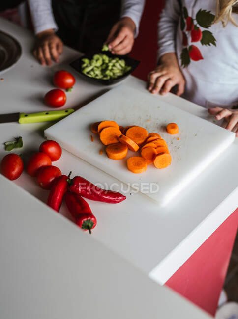 Вид на урожай анонімних маленьких дівчаток і хлопчика, які ріжуть і очищають стиглі овочі, готуючи здоровий салат на кухні разом — стокове фото