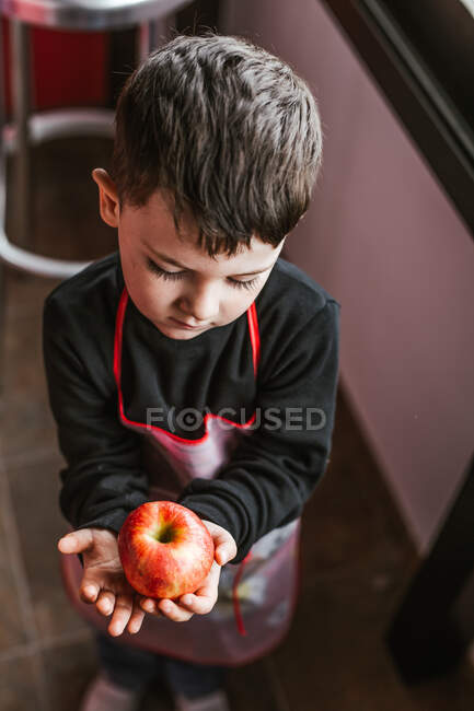 Мальчик показывает яблоко в камеру, стоя на кухне, глядя в камеру — стоковое фото