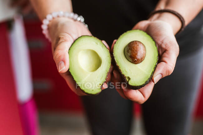 Неузнаваемая женщина демонстрирует две половинки спелого авокадо на камеру, стоя на размытом фоне на кухне — стоковое фото
