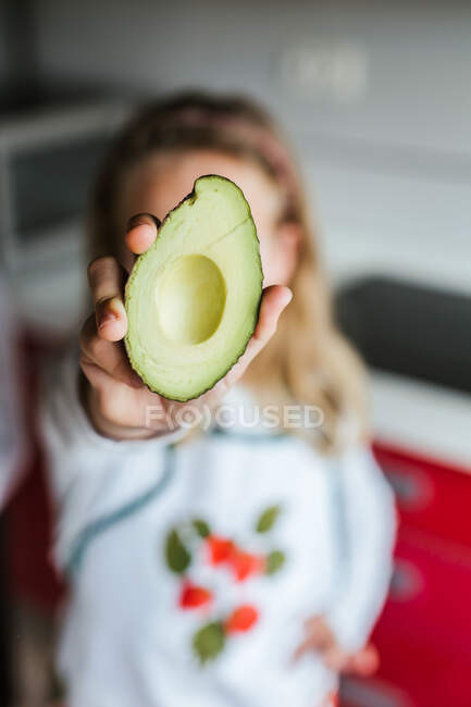 Неузнаваемая маленькая девочка демонстрирует половину спелого авокадо на камеру, стоя на размытом фоне на кухне — стоковое фото