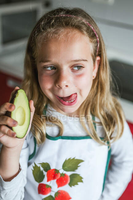 Kleines Mädchen mit der Hälfte der leckeren frischen Avocado schaut weg und leckt Lippen, während sie in der Küche steht — Stockfoto