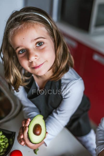 Маленька дівчинка демонструє половину стиглого авокадо до камери, стоячи на кухні, дивлячись на камеру — стокове фото
