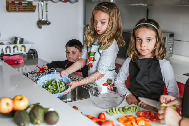 Niño y dos niñas en delantales de pie cerca del mostrador de la cocina y la preparación de verduras para la ensalada saludable en casa - foto de stock