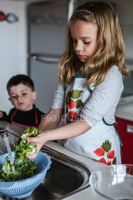 Little girl washing fresh herbs in sieve under clean water while making salad in kitchen — Stock Photo