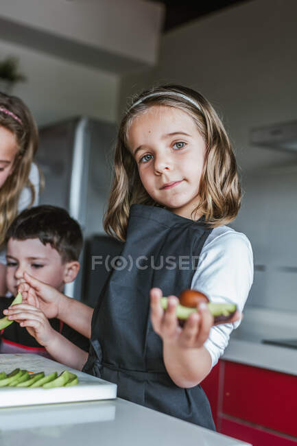 Маленькая девочка демонстрирует половину спелого авокадо на камеру, стоя на кухне и глядя в камеру — стоковое фото
