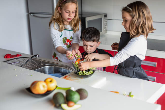 Little girls and boy cutting and peeling ripe vegetables while cooking healthy salad in kitchen together — Stock Photo