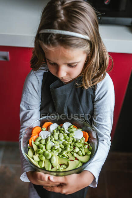 Kleines Mädchen mit Schale mit gesundem Gemüsesalat schaut in der heimischen Küche nach unten — Stockfoto