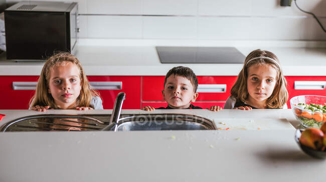Garçon et deux filles regardant du comptoir avec évier tout en jouant dans la cuisine à la maison ensemble — Photo de stock