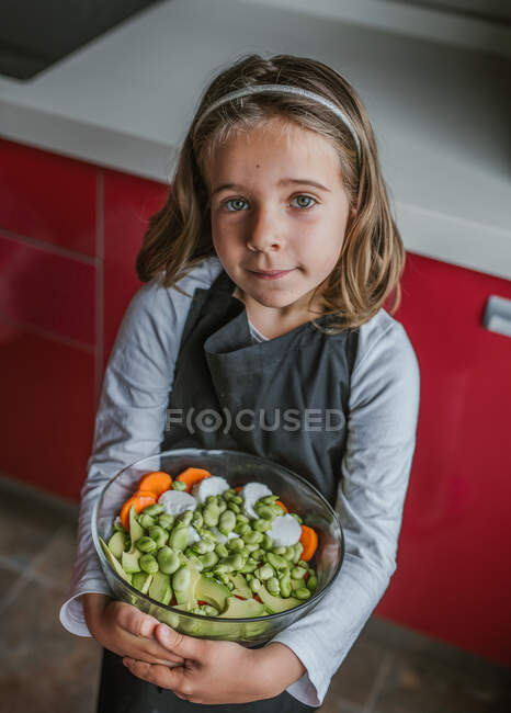 Petite fille avec bol de salade de légumes sains regardant la caméra tout en se tenant dans la cuisine à la maison — Photo de stock