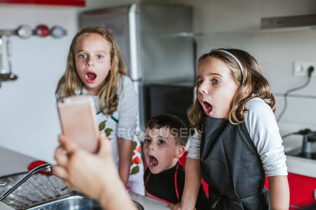Три вражені діти дивляться на екран смартфона в руці врожаю, стоячи на кухні разом — стокове фото