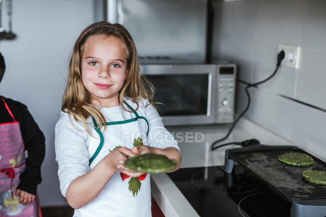 Petite fille souriant et regardant la caméra tout en tenant la plaque avec des escalopes végétariennes vertes dans la cuisine à la maison — Photo de stock