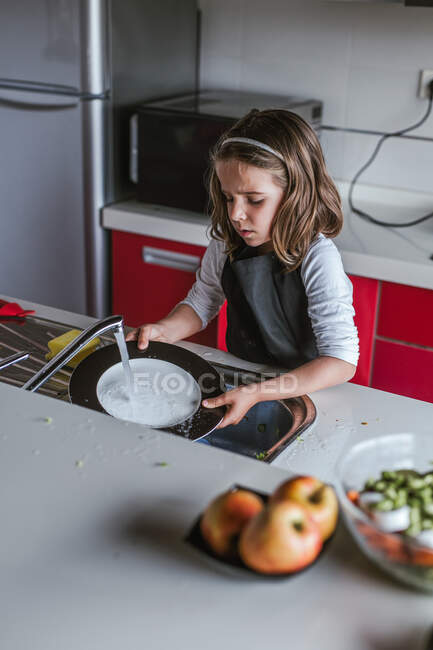 Маленькая девочка моет посуду над раковиной на кухне дома — стоковое фото