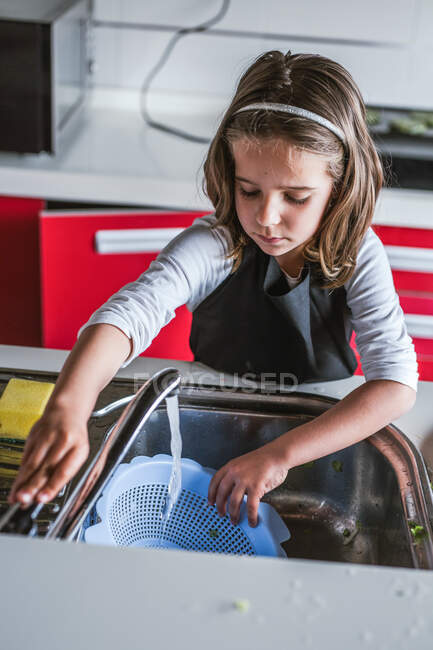 Маленькая девочка моет посуду над раковиной на кухне дома — стоковое фото