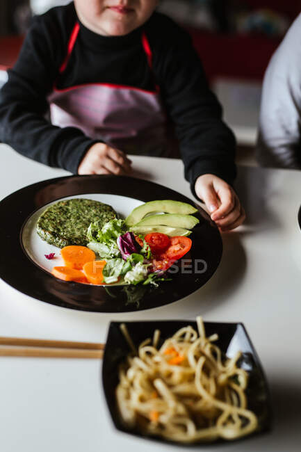 Niño anónimo esperando para comer sabrosos fideos con chuletas vegetarianas y verduras mientras está sentado en la mesa en casa - foto de stock