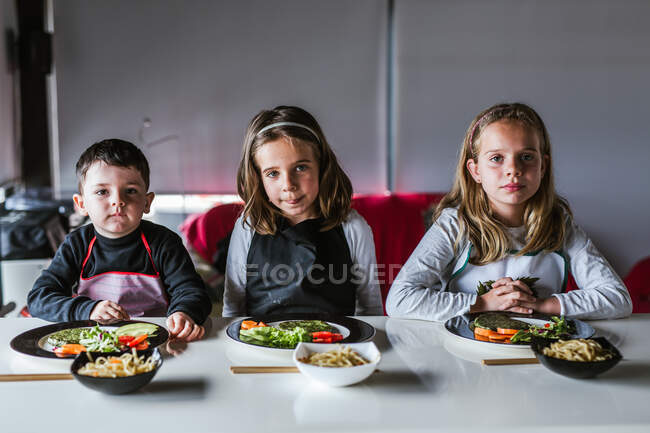 Junge und zwei Mädchen warten auf leckere Nudeln mit vegetarischen Schnitzeln und Gemüse, während sie zu Hause am Tisch sitzen — Stockfoto