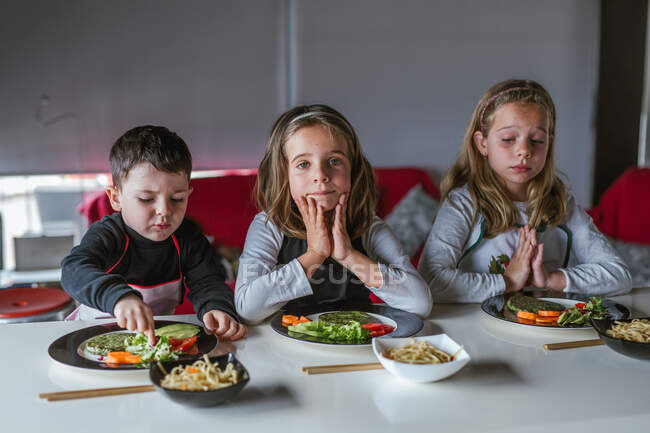 Хлопчик і дві дівчини чекають на смачну локшину з вегетаріанськими котлети і овочі, сидячи за столом вдома — стокове фото