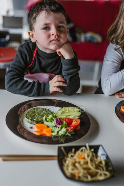 Niño esperando para comer sabrosos fideos con chuletas vegetarianas y verduras mientras está sentado en la mesa en casa - foto de stock