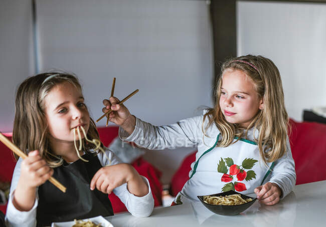 Две девушки едят вкусную лапшу с вегетарианскими котлетами и овощами, сидя за столом дома — стоковое фото