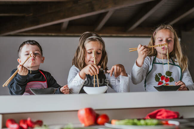 Ragazzo e due ragazze che mangiano gustosi spaghetti con costolette vegetariane e verdure mentre sono seduti a tavola a casa — Foto stock