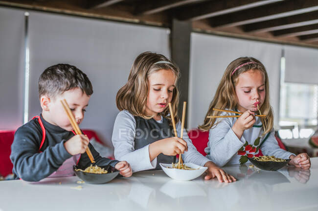 Хлопчик і дві дівчини їдять смачну локшину з вегетаріанськими котлети і овочі, сидячи за столом вдома — стокове фото