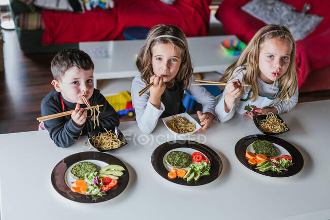 Ragazzo e due ragazze che guardano la macchina fotografica mentre si godono gustosi spaghetti con costolette vegetariane e verdure mentre sono seduti a tavola a casa — Foto stock