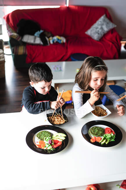 Garçon et deux filles dégustant des nouilles savoureuses avec des escalopes végétariennes et des légumes assis à la table à la maison — Photo de stock