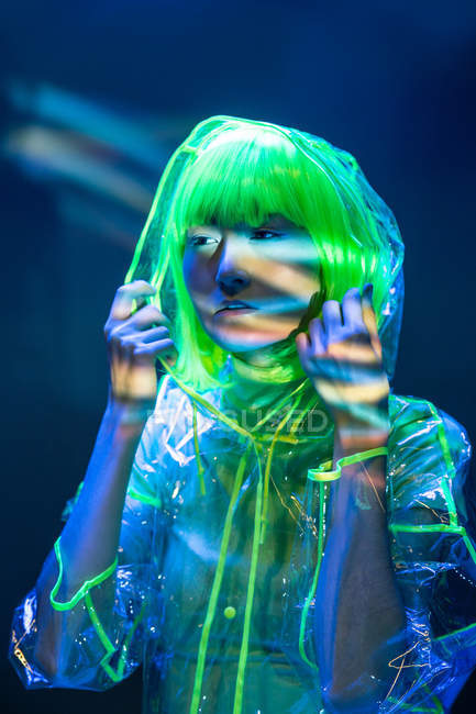 Jeune femme asiatique en perruque jaune et l'usure en plastique transparent posant dans la lumière fluorescente — Photo de stock