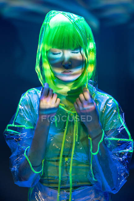 Giovane donna asiatica in parrucca gialla e plastica trasparente indossare posa in luce fluorescente — Foto stock