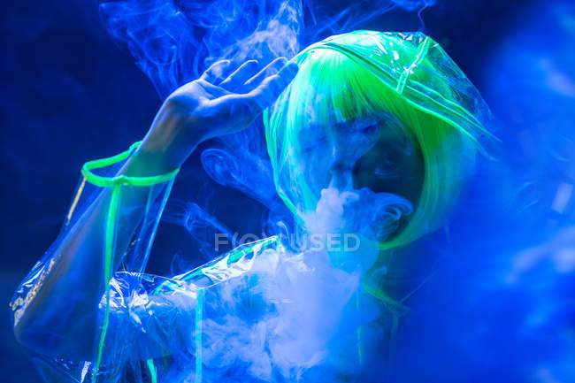 Giovane donna asiatica piuttosto insolita in plastica impermeabile trasparente e capelli gialli fumare in luce fluorescente — Foto stock