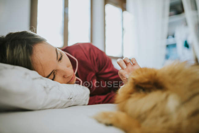 Молодая счастливая улыбающаяся привлекательная женщина в пижаме лежит в постели с маленькой пушистой собачкой — стоковое фото